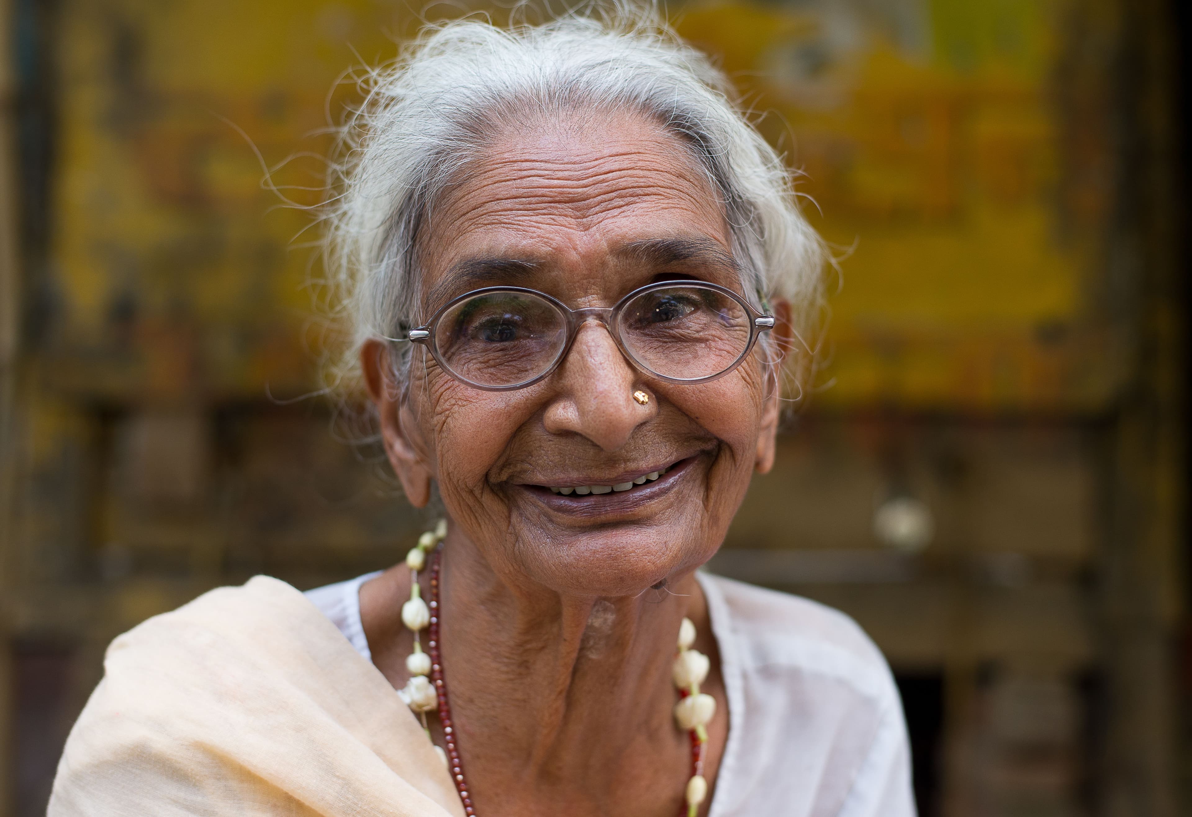 An elderly Indian woman with glasses smiling warmly outdoors